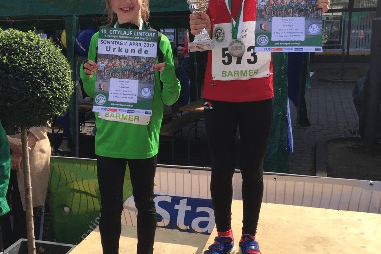 Zwei M&auml;dchen stehen auf Siegerpodesten, eines h&auml;lt eine Urkunde und das andere einen Pokal beim Citylauf in Lintorf.