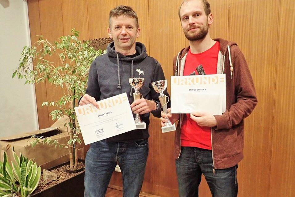 Zwei M&auml;nner halten Pokale und Urkunden in der Hand, umgeben von Pflanzen in einem Innenraum.