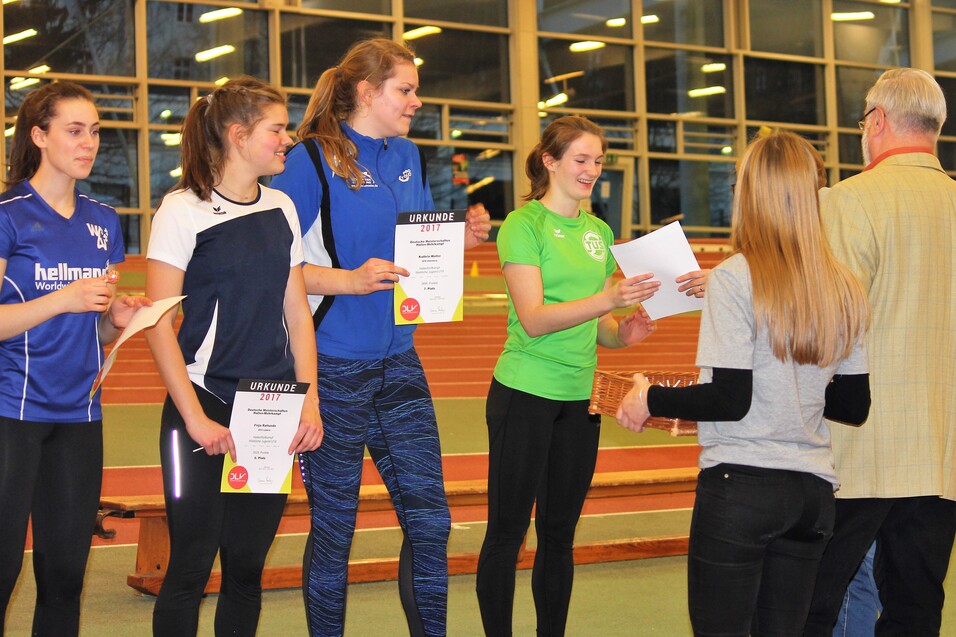 Zwei Gruppen von Personen stehen in einer Sporthalle; eine Person &uuml;berreicht Urkunden an Sportlerinnen.