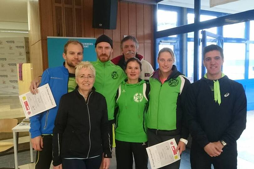 Gruppenfoto von sieben Personen in sportlicher Kleidung, die Urkunden halten, vor einer Fensterfront in einem Gebäude.