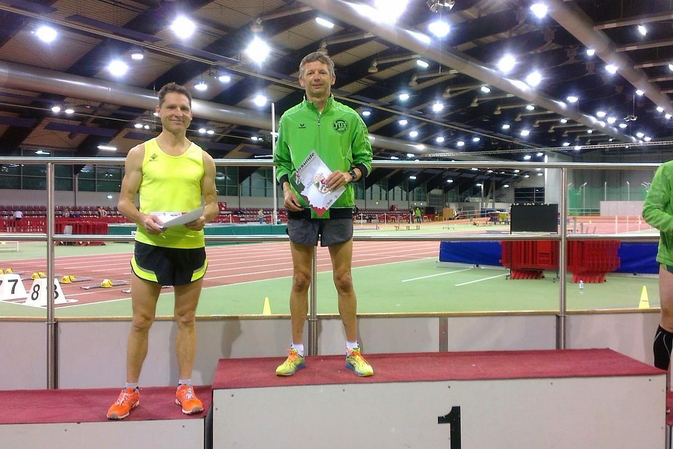 Zwei Athleten stehen auf einem Siegerpodest in Sportanz&uuml;gen, der erste h&auml;lt ein Zertifikat in der Hand.
