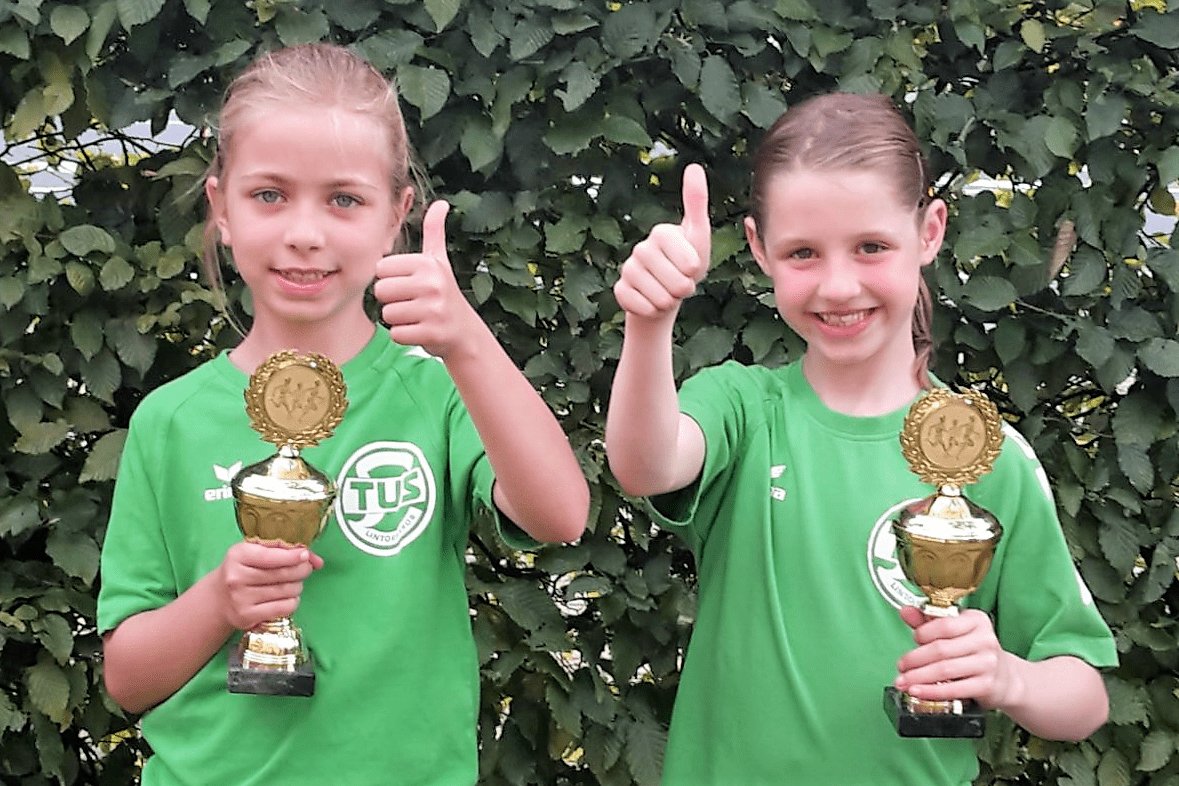 Zwei M&auml;dchen in gr&uuml;nen T-Shirts halten Pokale und zeigen Daumen hoch vor einer Hecke.