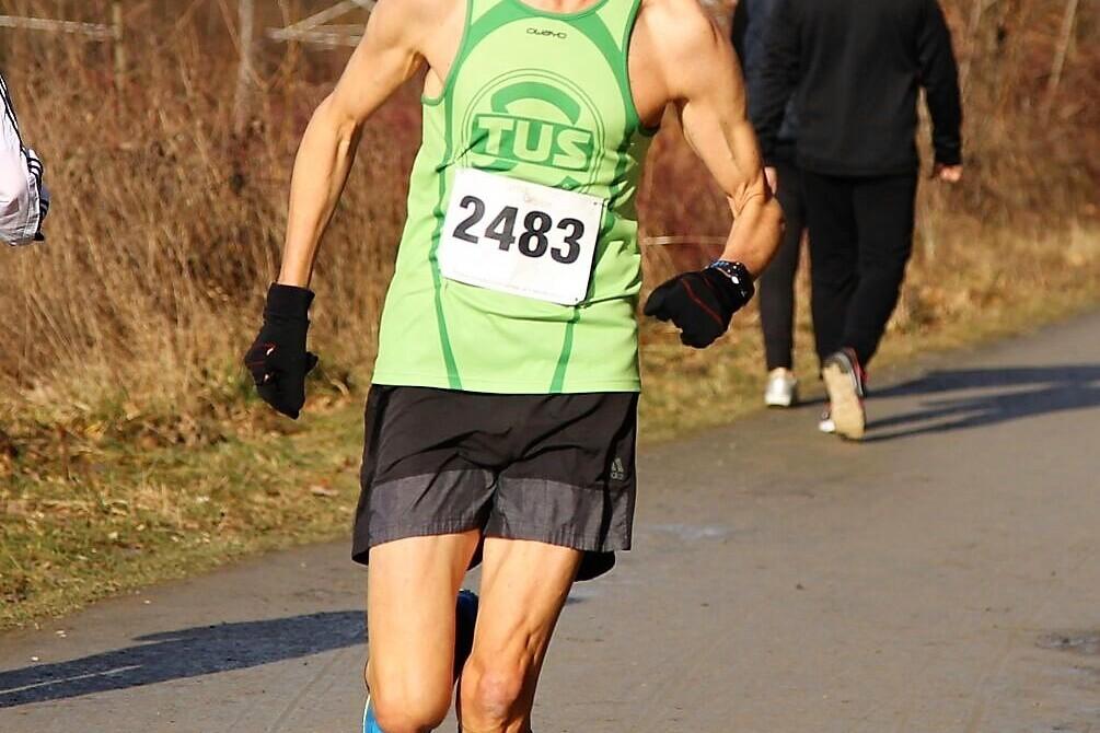 Läufer in grünem Trikot mit der Nummer 2483, der auf einem asphaltieren Weg unterwegs ist, umgeben von Bäumen.
