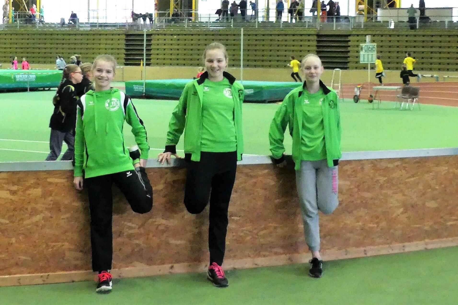 Drei sportliche M&auml;dchen in gr&uuml;ner Sportkleidung lehnen an einer Holzbarriere in einer Sporthalle.
