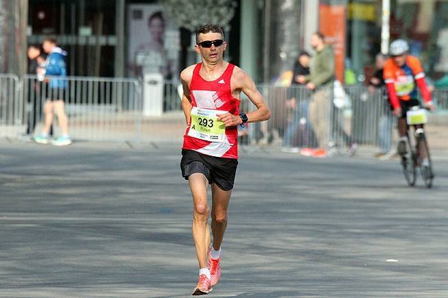 Läufer in rotem Trikot mit der Startnummer 293 joggt auf einer Straße, im Hintergrund stehen Zuschauer und ein Radfahrer.