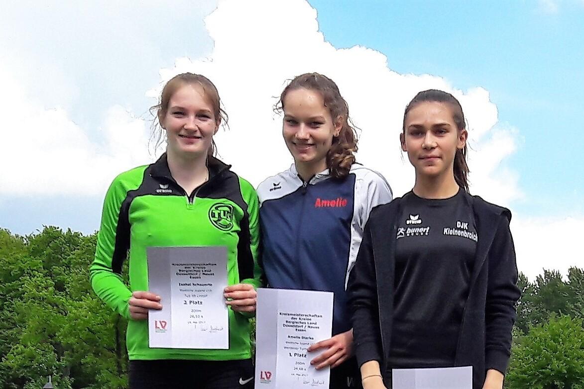 Drei junge Sportlerinnen stehen lächelnd mit Urkunden in der Hand vor einer grünen Landschaft.