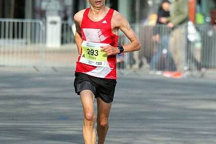 L&auml;ufer in rotem Trikot mit der Startnummer 293 joggt auf einer Stra&szlig;e, im Hintergrund stehen Zuschauer und ein Radfahrer.