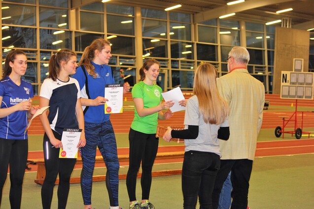 Zwei Gruppen von Personen stehen in einer Sporthalle; eine Person &uuml;berreicht Urkunden an Sportlerinnen.