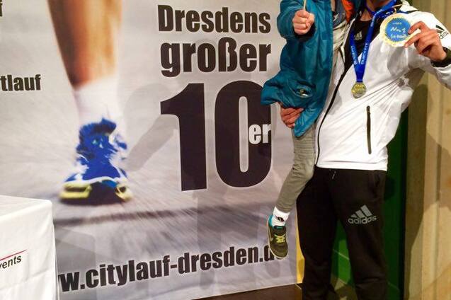 Mann mit Medaille h&auml;lt l&auml;chelnd ein Kind auf dem Arm vor einem Banner des Citylaufs Dresden.