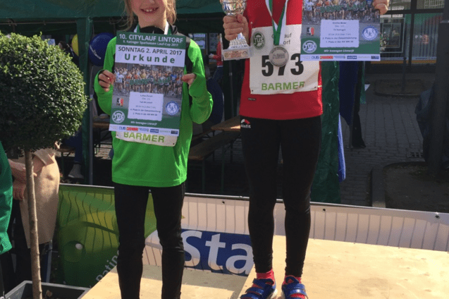 Zwei M&auml;dchen stehen auf Siegerpodesten, eines h&auml;lt eine Urkunde und das andere einen Pokal beim Citylauf in Lintorf.