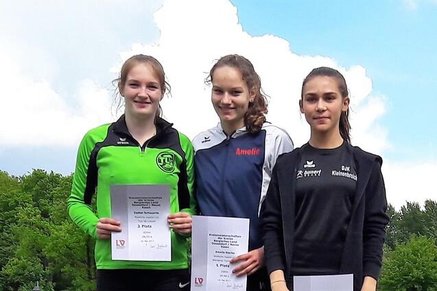 Drei junge Sportlerinnen stehen l&auml;chelnd mit Urkunden in der Hand vor einer gr&uuml;nen Landschaft.