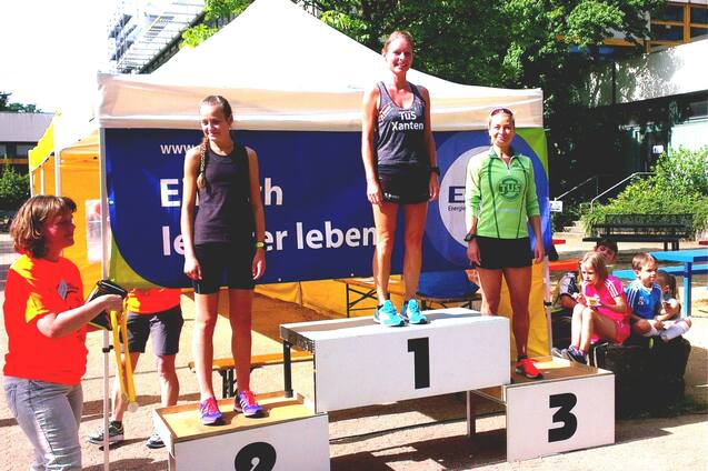 Drei Frauen auf dem Siegerpodest eines Wettkampfs, mit Platz 1 in der Mitte und Zuschauern im Hintergrund.