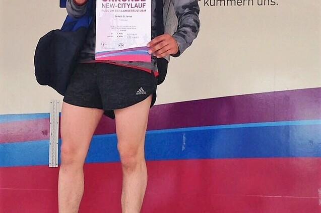 Mann steht auf dem ersten Siegerpodest und h&auml;lt die Urkunde des New-Citylaufs in der Hand, l&auml;chelt und tr&auml;gt Sportkleidung.