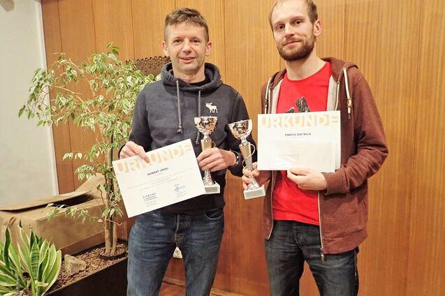 Zwei M&auml;nner halten Pokale und Urkunden in der Hand, umgeben von Pflanzen in einem Innenraum.