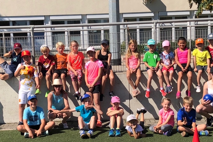 Gruppe von Kindern in Sportbekleidung sitzt und steht auf einer Mauer und einem Fu&szlig;ballfeld bei sonnigem Wetter.