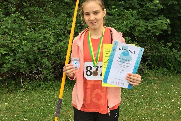 M&auml;dchen mit einer Medaille und Urkunde h&auml;lt einen Speer in der Hand, vor gr&uuml;ner Kulisse im Freien.
