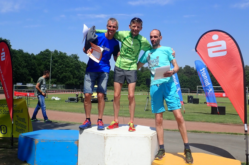 Drei L&auml;ufer stehen auf dem Siegertreppchen, umgeben von Flaggen und einer Sportanlage im Hintergrund.