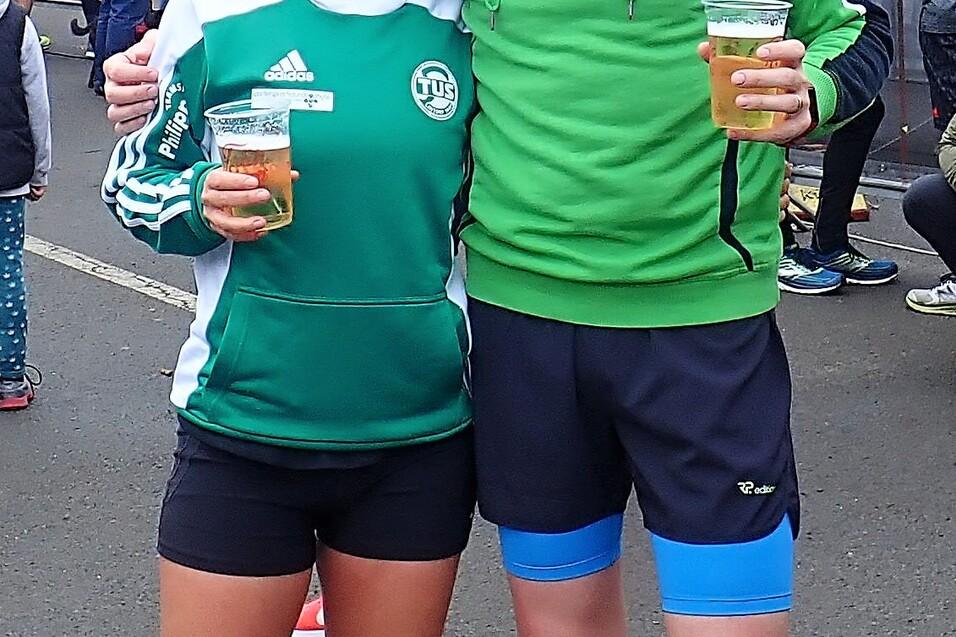 Zwei L&auml;ufer halten Bier in der Hand, umgeben von einer Menschenmenge und bunten Werbefahnen bei einer Veranstaltung.