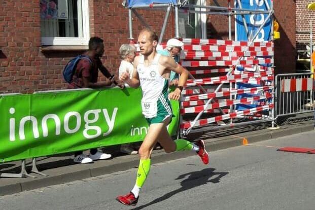 Läufer in Sportkleidung sprintet bei einem Wettbewerb vor Zuschauern und einer grünen Werbebande entlang der Straße.