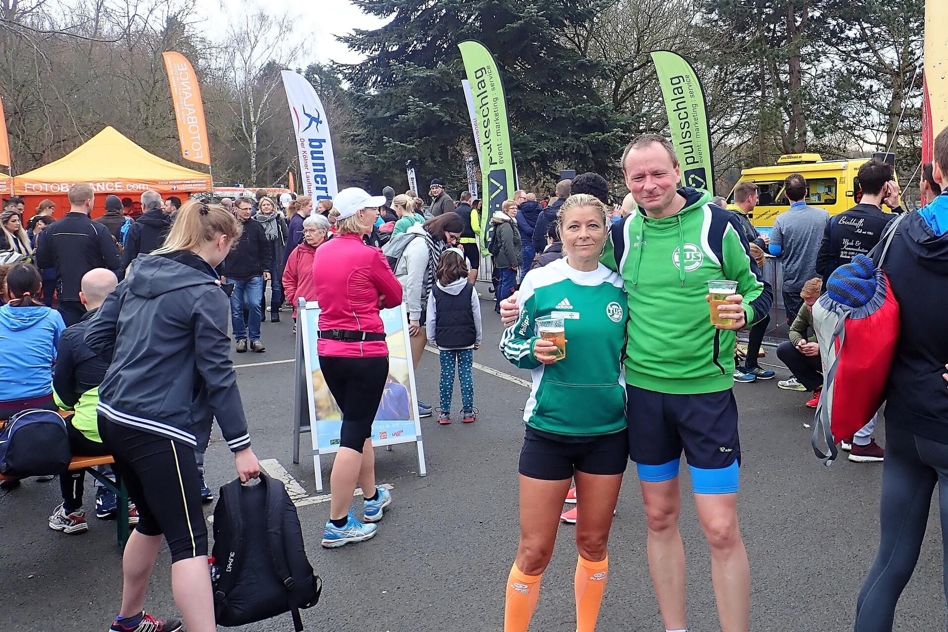 Zwei L&auml;ufer halten Bier in der Hand, umgeben von einer Menschenmenge und bunten Werbefahnen bei einer Veranstaltung.