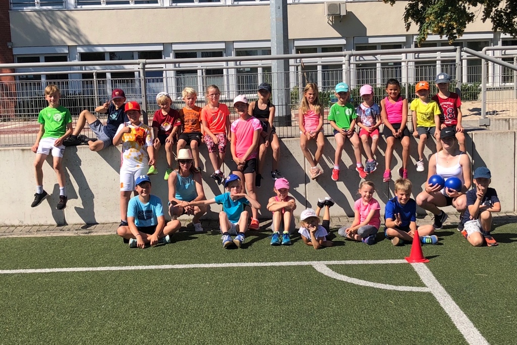 Gruppe von Kindern in Sportbekleidung sitzt und steht auf einer Mauer und einem Fu&szlig;ballfeld bei sonnigem Wetter.