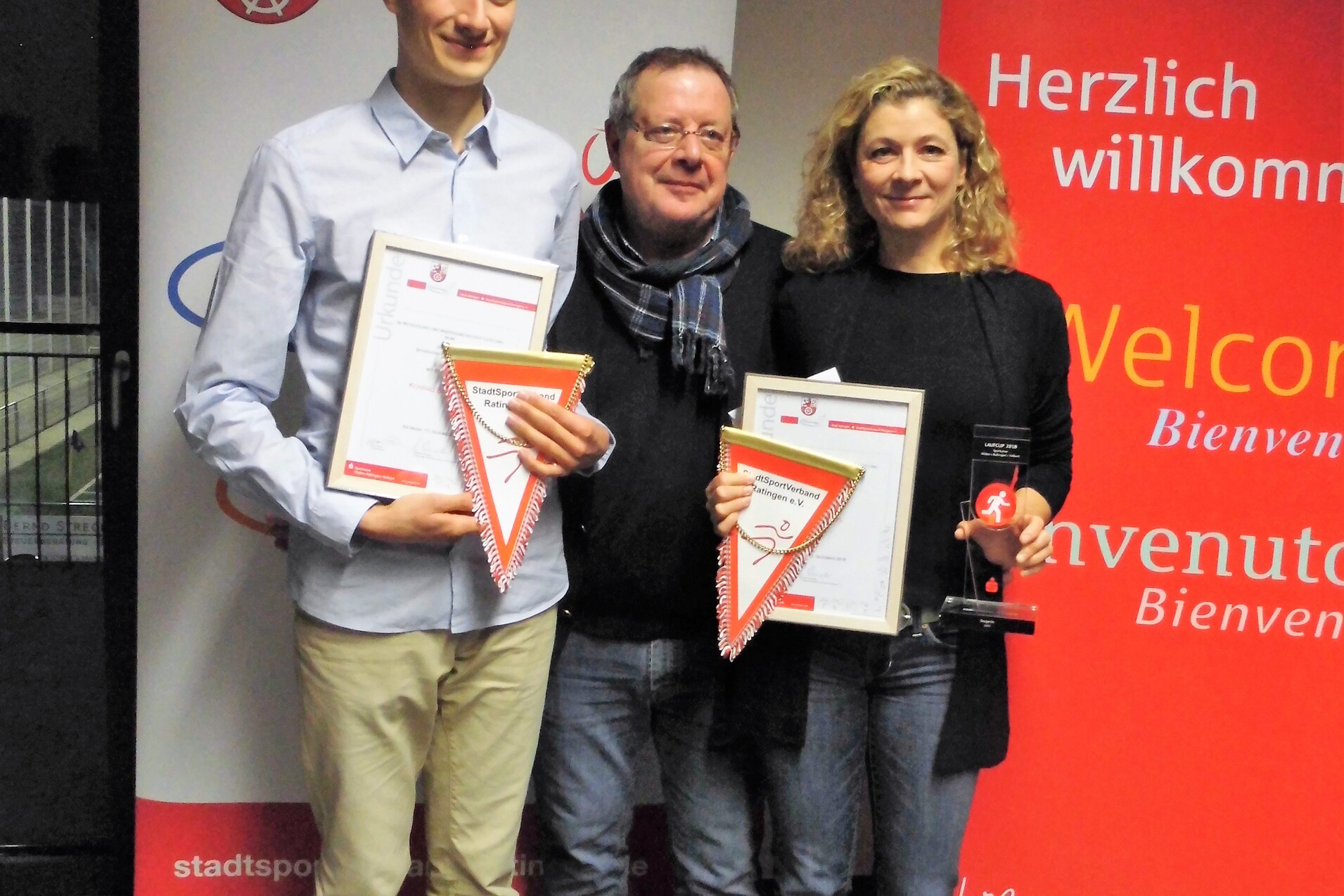 Drei Personen halten Urkunden in der Hand, vor einem Banner mit Willkommenstext und Logos im Hintergrund.