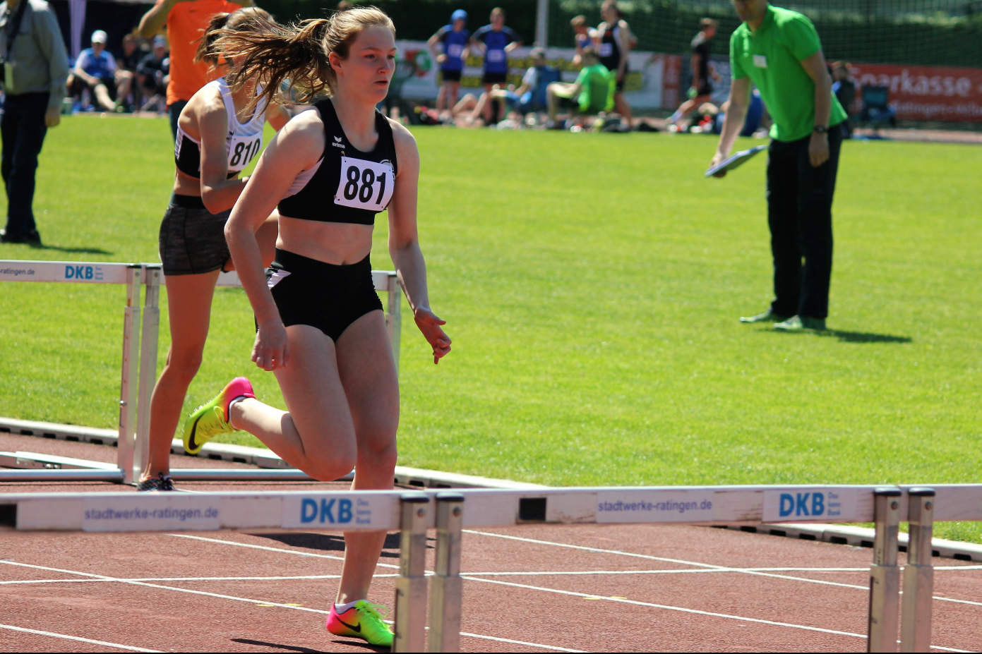 Athletin mit Startnummer 881 &uuml;berquert die H&uuml;rden auf einem Sportplatz, im Hintergrund Zuschauer und eine gr&uuml;ne Rasenfl&auml;che.