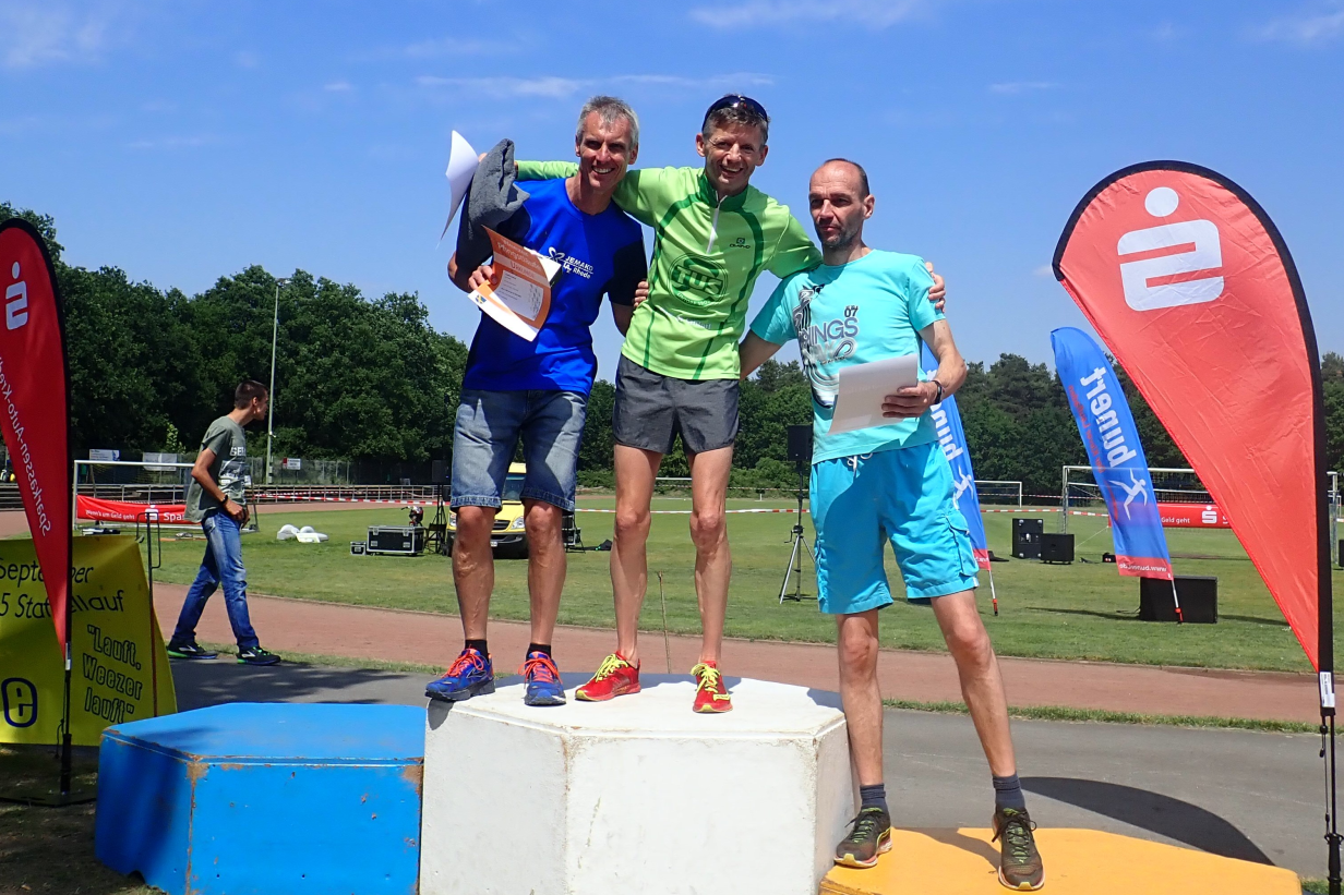 Drei L&auml;ufer stehen auf dem Siegertreppchen, umgeben von Flaggen und einer Sportanlage im Hintergrund.
