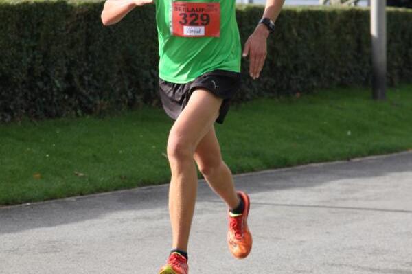 L&auml;ufer in gr&uuml;nem T-Shirt sprintet auf einem B&uuml;rgersteig w&auml;hrend eines Wettkampfs, umgeben von zuschauenden Menschen.