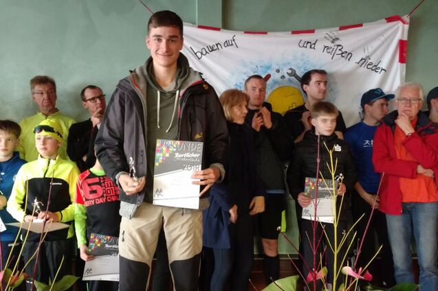 Junger Mann mit Urkunde steht vor einer Gruppe von Menschen bei einer Veranstaltung, mit einer dekorativen Banner im Hintergrund.