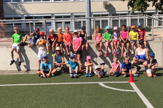 Gruppe von Kindern in Sportbekleidung sitzt und steht auf einer Mauer und einem Fu&szlig;ballfeld bei sonnigem Wetter.