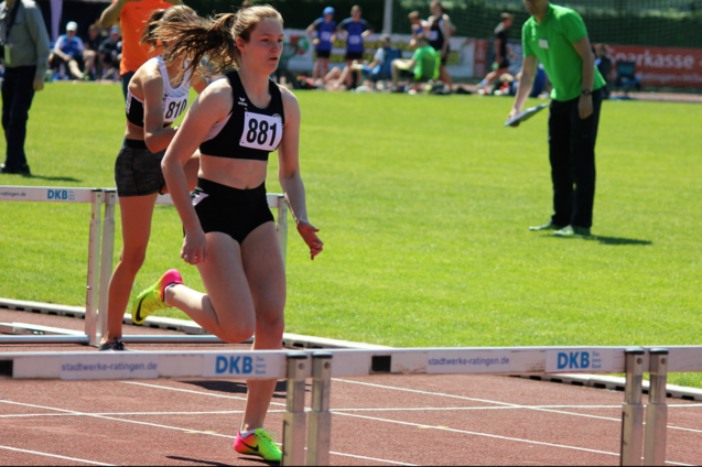 Athletin mit Startnummer 881 &uuml;berquert die H&uuml;rden auf einem Sportplatz, im Hintergrund Zuschauer und eine gr&uuml;ne Rasenfl&auml;che.
