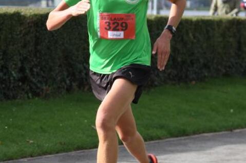 L&auml;ufer in gr&uuml;nem T-Shirt sprintet auf einem B&uuml;rgersteig w&auml;hrend eines Wettkampfs, umgeben von zuschauenden Menschen.