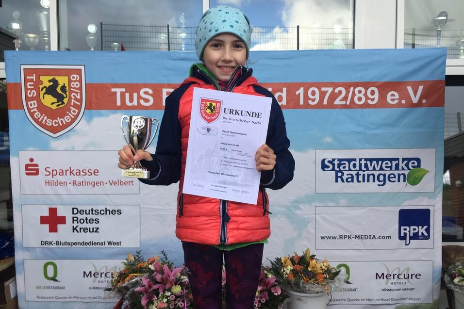 Ein M&auml;dchen h&auml;lt eine Urkunde und einen Pokal vor einer Ehrenwand mit Logos und Blumenschmuck.