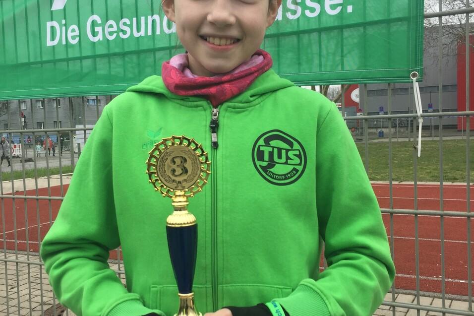 M&auml;dchen in gr&uuml;ner Jacke h&auml;lt einen Pokal vor einer Bannerwand mit dem AOK-Logo.