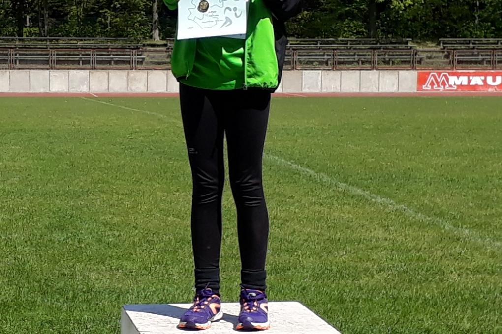 Mädchen steht auf dem ersten Platz eines Sportpodests und hält eine Urkunde in der Hand. Sportplatz im Hintergrund.