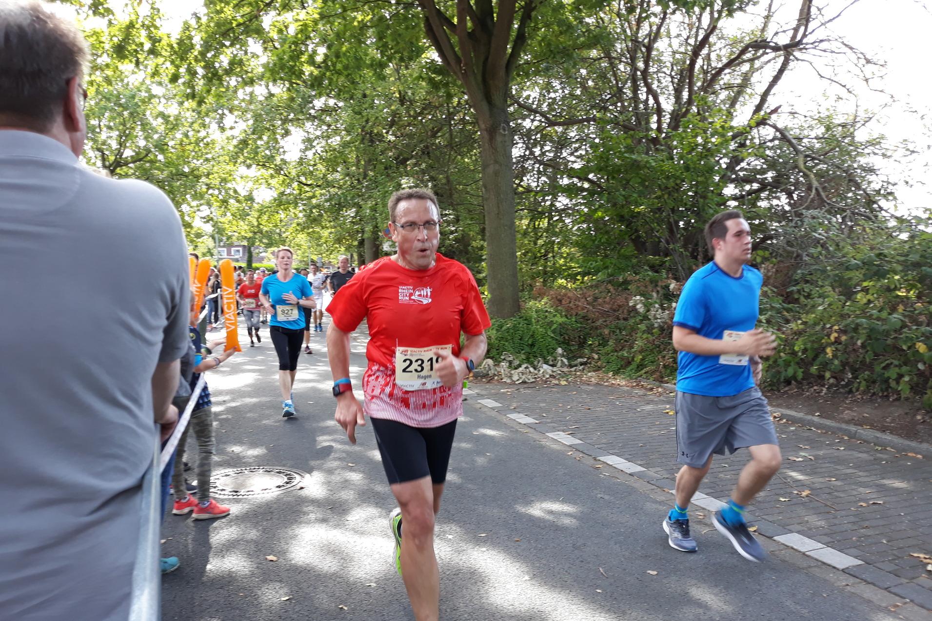 L&auml;ufer in roten und blauen Shirts bei einem Stra&szlig;enlauf, umgeben von B&auml;umen und Zuschauern.