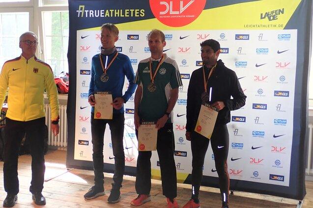 Drei Athleten mit Medaillen und Urkunden stehen vor einer Sponsorenwand bei einer Preisverleihung.