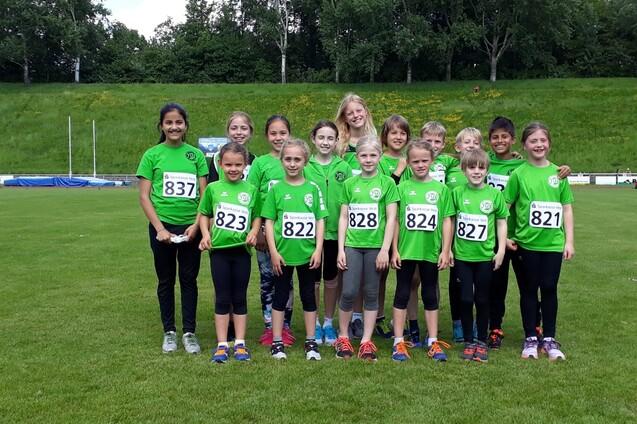 Gruppe von Jugendlichen in gr&uuml;nen T-Shirts mit Startnummern, stehen auf einem Sportplatz im Freien.