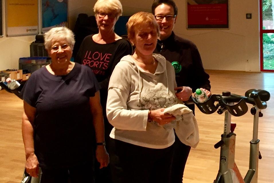 Gruppe &auml;lterer Frauen posiert l&auml;chelnd vor Fitness-Bikes in einem Trainingsraum mit Fenstern.