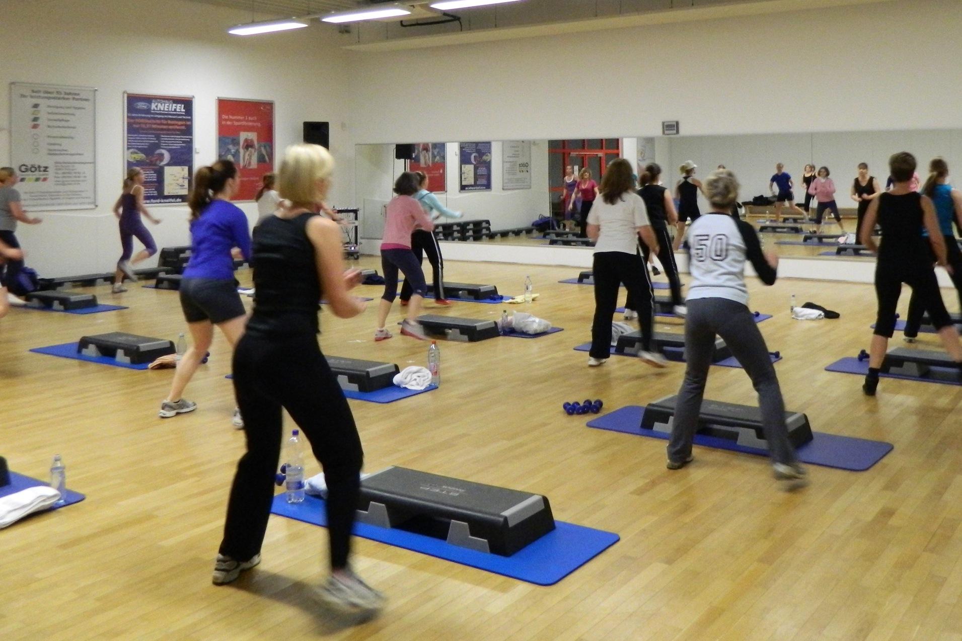Gruppe von Frauen bei einem Fitnesskurs in einem Spiegelraum, mit Step-Brettern und Gewichten auf dem Boden.
