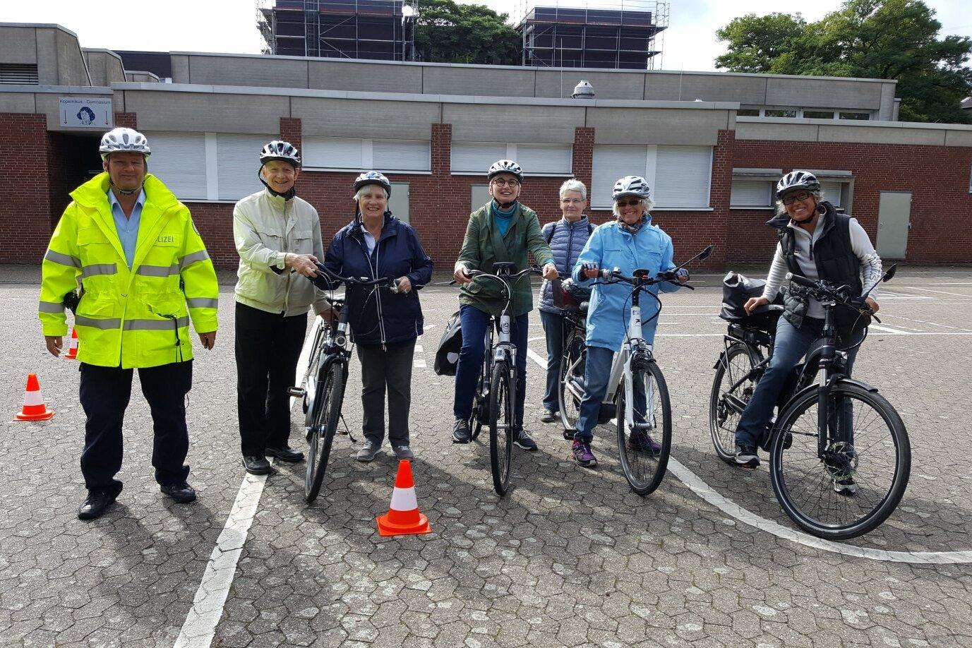 Gruppe von sechs Personen mit Fahrradhelmen steht mit Fahrr&auml;dern auf einem markierten Platz, daneben ein Helfer in Warnjacke.
