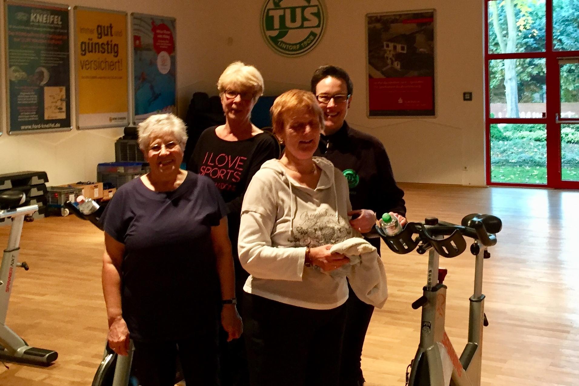 Gruppe &auml;lterer Frauen posiert l&auml;chelnd vor Fitness-Bikes in einem Trainingsraum mit Fenstern.