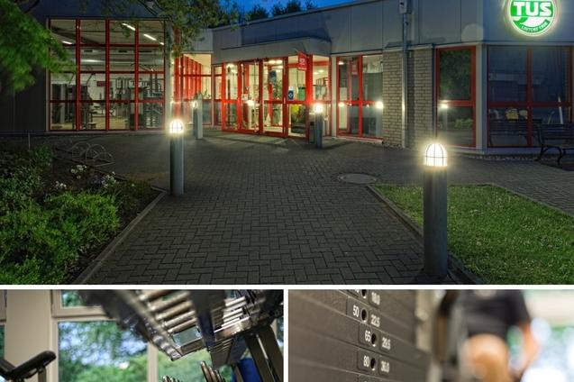 Eingang eines Fitnessstudios bei D&auml;mmerung mit beleuchtetem Logo und Umgebungsbeleuchtung, darunter Fitnessger&auml;te und Gewichte.