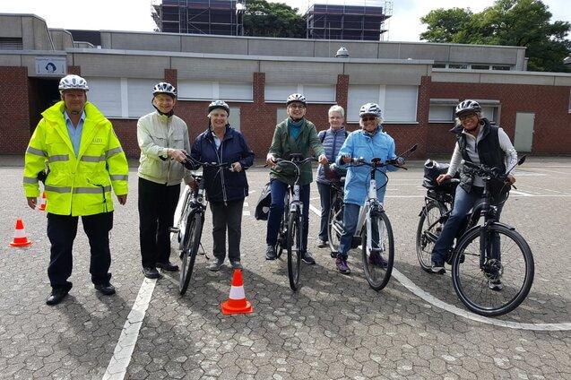 Gruppe von sechs Personen mit Fahrradhelmen steht mit Fahrr&auml;dern auf einem markierten Platz, daneben ein Helfer in Warnjacke.