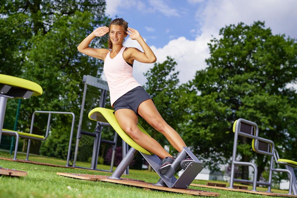 Frau in Sportkleidung macht seitliche Rumpfbeuge auf Trainingsger&auml;t im Freien, begr&uuml;nte Umgebung im Hintergrund.