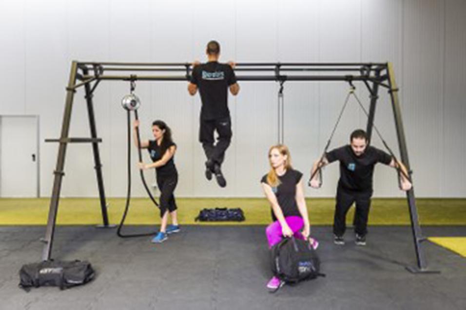 Vier Personen trainieren an einer Fitnessstation mit einem Trainingsger&auml;t, Gewichten und Fitness-Taschen.