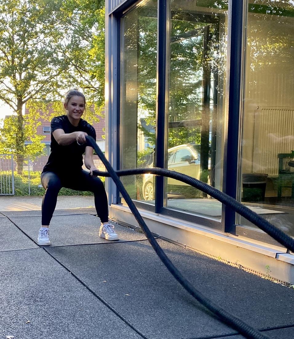 Frau in Sportkleidung trainiert mit Fitnessseilen auf einem Outdoor-Bereich vor gro&szlig;en Fenstern.