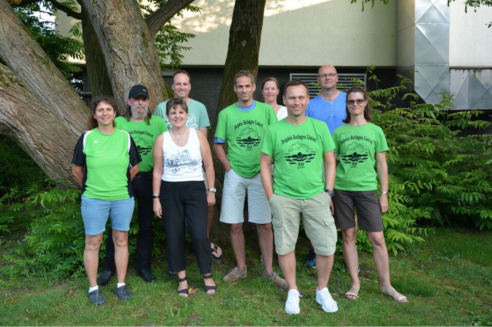 Gruppe von acht Personen in gr&uuml;nen T-Shirts steht unter einem Baum in einer lichtdurchfluteten Umgebung.