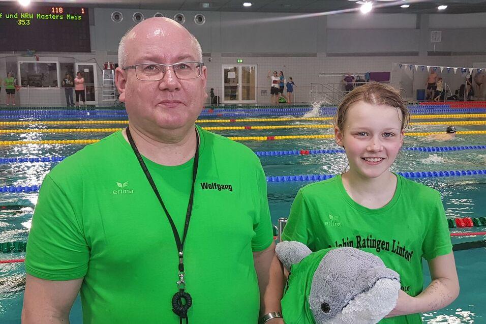 Ein Trainer und ein M&auml;dchen in gr&uuml;nen T-Shirts stehen am Beckenrand mit einem Pl&uuml;schdelfin in der Hand.